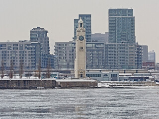 Obraz premium Montreal clock tower skyscrapers and industrial buildings on Quai d`horloge in the Old Port of Montreal, view from Jean Drapeau park across Saint Lawrence river on a hazy winter day. Quebec, Canada 