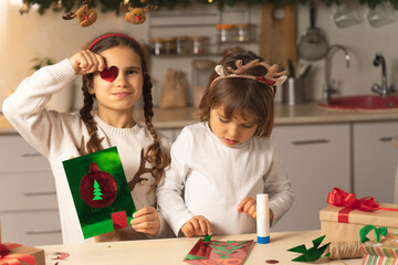 Cheerful Kids make a Christmas card for mom. Funny sisters prepare for Christmas and New Year at home in the kitchen.