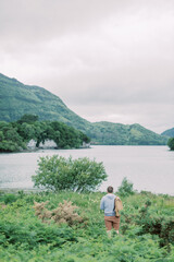 A young man stands at lake looking at mountains ahead in Ireland