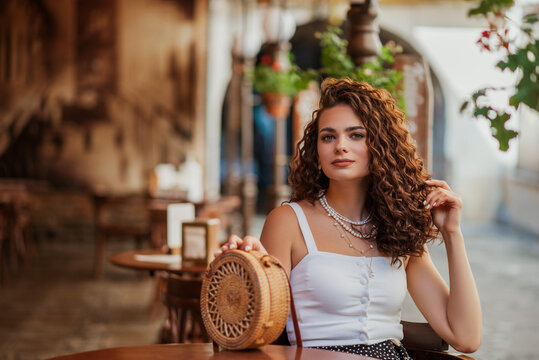 Fototapeta Elegant fashionable curly brunette woman posing in street cafe in European city. Summer fashion, vacation, travel, lifestyle conception. Copy, empty space for text