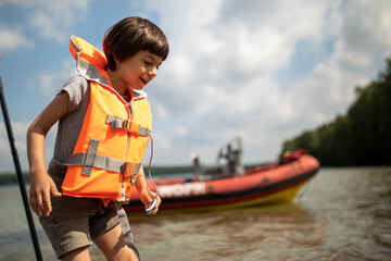 Little adorable boy in orange life vest stepping in water of lake in summer