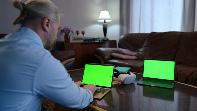 Two chromakey laptops with web page mockup and Caucasian man typing messaging online. Shooting over shoulder of confident successful startuper surfing internet in home office indoors. Template