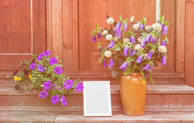 Flower pot with beautiful petunias, empty white frame  and clay vase with bellflowers on steps of rustic wooden ladder.