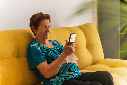 Older Caucasian Woman Using Mobile Phone Sitting On Yellow Sofa. She Is Wearing A Blue T-shirt, Natural Light Is Coming In.