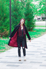 A young woman with long hair and a red hat wearing glasses and dark coat with a red scarf in the park walking against trees on a pedestrian road.