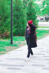 A young woman with long hair and a red hat wearing glasses and dark coat with a red scarf in the park walking against trees on a pedestrian road.
