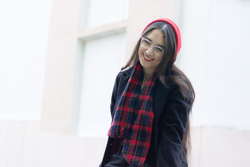 Fototapeta premium A headshot of a young woman wearing a red hat, red scarf and a black coat is sitting on a green fance against the white and orange classic building at the street