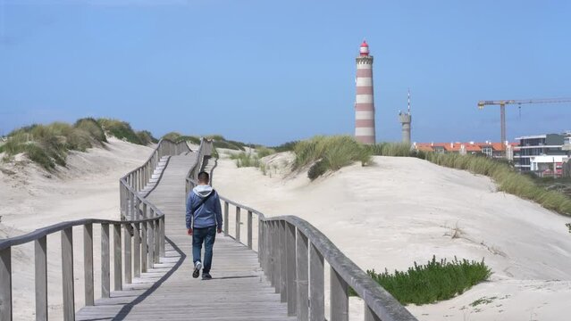 Man walking at wooden walkway at beach. Sand dunes against lighthouse, Praia da Barra, Aveiro