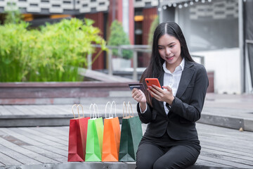 Happy young asian woman holding credit card and smartphone for online shopping in park.