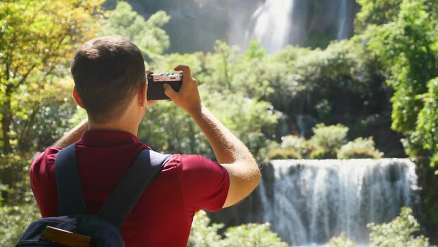 Young man traveler make photos for social media of beautiful exotic waterfalls and green tropical nature on retro style camera. Happy male tourist with backpack in travel. Wanderlust, tourism concept.