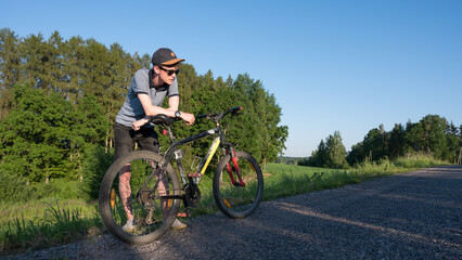 Fototapeta premium A young man with a bicycle is standing on the edge of the road. Sunny summer. Green trees. Countryside.