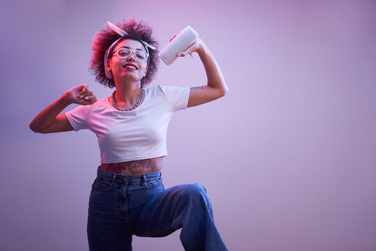 Young positive kazakh girl with afro hairstyle listens music in portable speaker, dances and poses, hip hop style