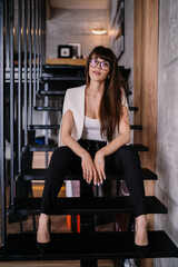 Fototapeta na wymiar Young Italian brunette with loose hair dressed elegantly sitting on the steps in office, needs a brake. Vertical shot of beautiful caucasian girl in glasses exhausted after work. Lawyer at office