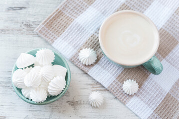 Small meringue in the ceramic bowl and cup of coffee with cream on the kitchen towel on the rustic background. Flat lay