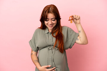 Young redhead caucasian woman isolated on pink background pregnant and holding a pacifier