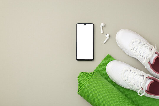 Fitness accessories concept. Top view photo of smartphone earbuds white sneakers and green sports mat on isolated pastel grey background with blank space