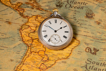 Silver retro pocket watch lying on a paper old world map. Antique round clock with a dial and arrows on a geographical map with continents and sea. The concept of time, travel, adventure, treasure.