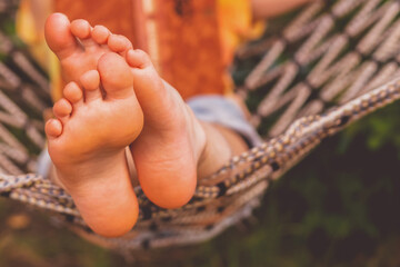 Young beautiful girl lies in a hammock and reads a book. Rest, summer vacation, leisure time concept. Selective focus on feets. Copy space.