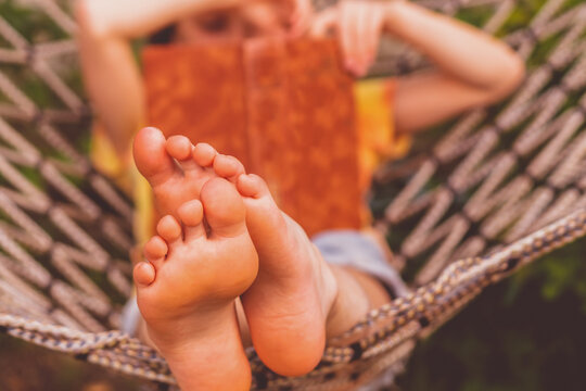 A young beautiful girl lies in a hammock and reads a book. Rest, summer vacation, leisure time concept. Selective focus on feets. Horizontal image.