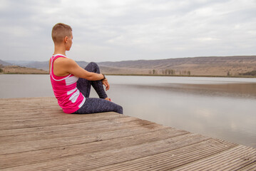 young woman with short hair in sportswear sitting on the wooden bridge near lake in the mountains