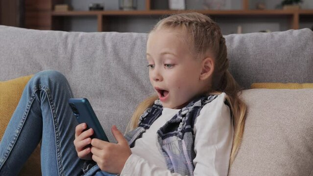 Portrait of surprised little cute girl holding smartphone lying at sofa at home. Choosing favorite music or cartoons,texting messages,browsing internet,watching video, playing games on smartphone