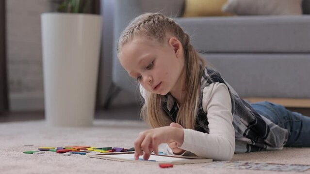 Educational toys for children. Cute little girl making a wooden puzzle with colorful blocks lying on the carpet at home. Brain teasers toy for motor development and logical thinking. Home leisure.