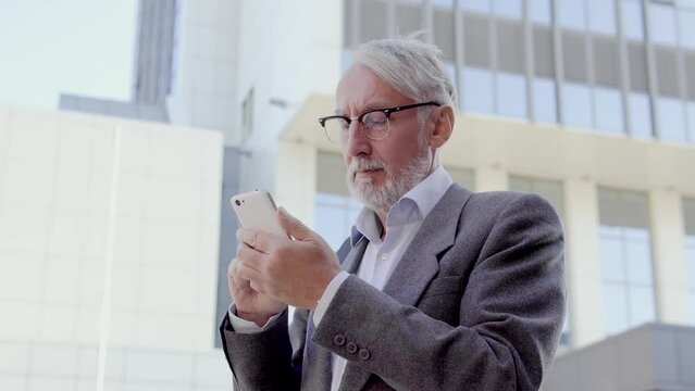Successful gray-haired man typing message on smartphone standing outdoor, app