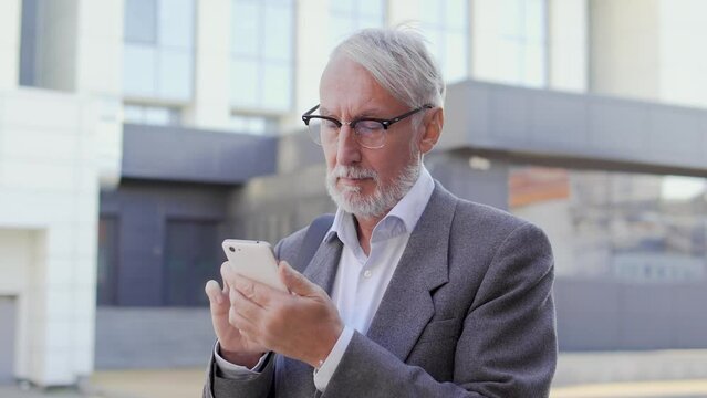 Busy bearded man in stylish eyeglasses and business suit scrolling on smartphone standing outdoor, reading and applying emails, checking news and stocks market rates, free wi-fi