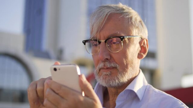 Handsome mature man in eyeglasses scrolling web-pages on smartphone standing outdoor, chatting in messenger or in dating app with friends, ordering delivery, digitalization, close-up