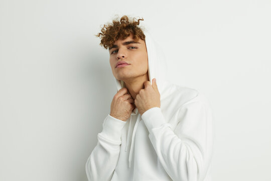 a cute man stands on a white background in a white hoodie and looks pleasantly into the camera adjusting the hood with his hands