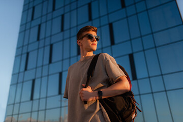 Portrait of handsome tourist hipster man in sunglasses with bag handy, standing guy near the building in downtown. Calm relaxing moment, summer vacation concept - 517065124