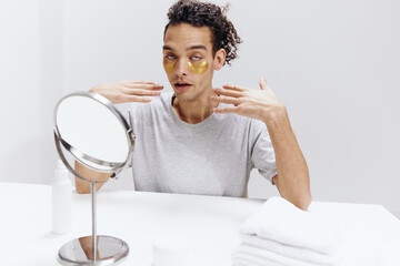guy with curly hair in a white T-shirt sits at a table with a mirror and a towel face patches light background