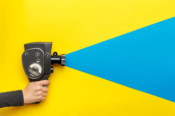 Female hand holding old style film movie camera on a yellow background with blue ray coming out of the camera,  imitating shooting process. - 517067532