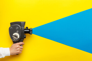 Female hand holding old style film movie camera on a yellow background with blue ray coming out of...