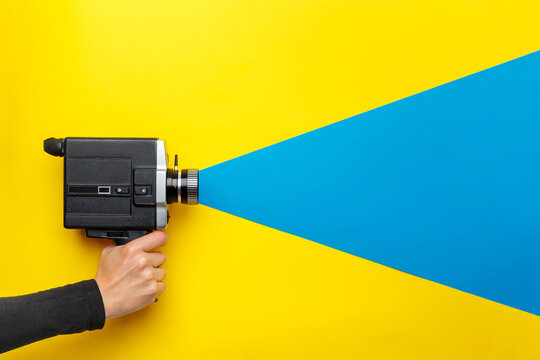 Female hand holding old style film movie camera on a yellow background with blue ray coming out of the camera,  imitating shooting process.