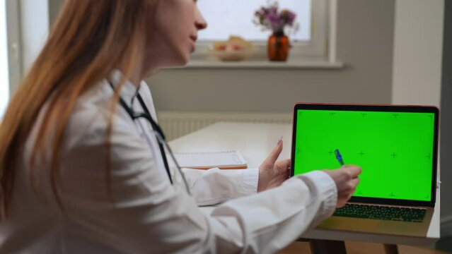 Unrecognizable blurred doctor pointing at green screen on laptop talking advertising web page mockup. Young slim Caucasian woman promoting treatment online site in hospital indoors