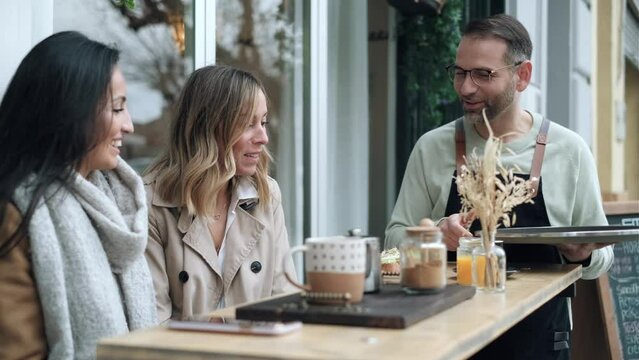 Video of handsome waiter serving toast for brunch to two modern girls sitting on the terrace on the healthy coffee shop terrace