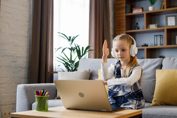 Primary schoolgirl wearing earphones has online lesson, video call with teacher, raising hand to answer the question. Cute child studying at home using laptop. Distance learning. Home education