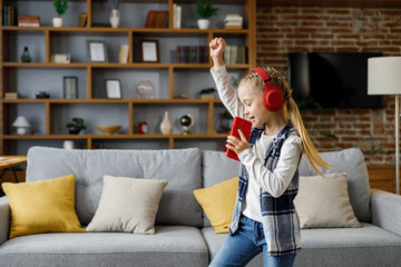 Happy little girl wearing red headphones dancing and singing using smartphone like microphone. Cute child enjoy listening favorite music tracks in mobile application Home leisure, happy childhood