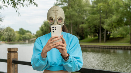 A girl in a gas mask takes a photo on the phone on the river bank. Alienation in the city. Fashion,...