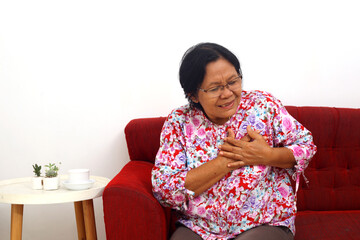 Senior adult woman sitting on the sofa while holding her chest having heart attack. Isolated