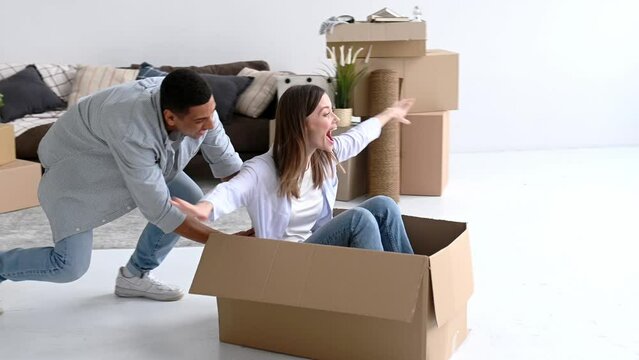 Couple in love, young man and woman of different nationalities, moved to their new home, fooling around with cardboard boxes, girl sits in a box, guy rolls her around the living room, laugh, have fun