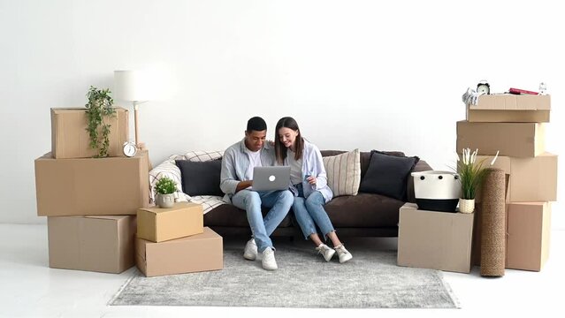 Real estate concept. Happy mixed race family couple sitting on a sofa between cardboard boxes in living room of their new home, using laptop and credit card for online shopping, buying things for home