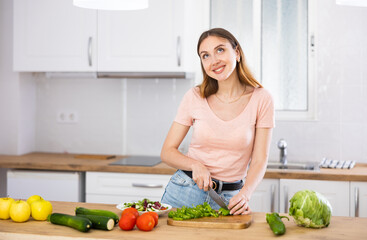Portrait of positive woman standing at kitchen table at home, slicing vegetables, cooking salad.