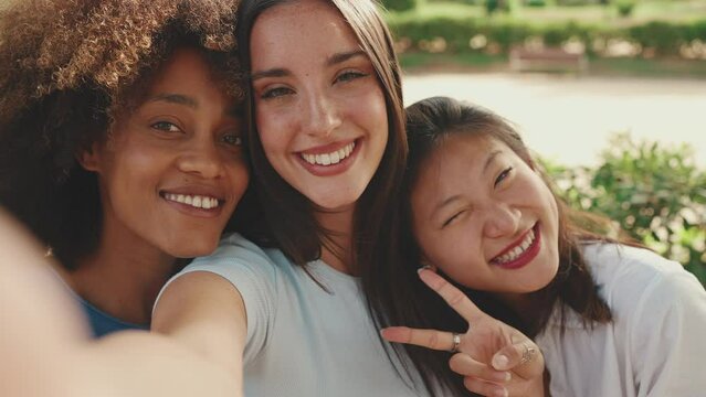 Happy, lovely multiethnic young women posing on cellphone camera on summer day outdoors. group of friends hugging each other smiling at the camera while standing on path in the park