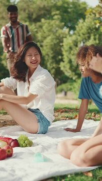 VERTICAL VIDEO: Happy smiling young multinational people at picnic on summer day outdoors. Guy arriving at picnic in the park, hugging, having fun spending the weekend together, relaxing