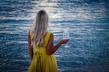 A blonde girl in a yellow dress is standing on the ocean. Glass of wine in hand. Soft focus © VK