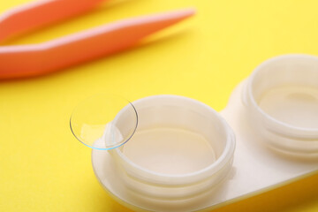 Case with contact lens and tweezers on yellow background, closeup