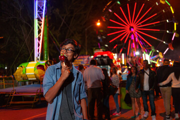 Happy Latino boy eating a caramelized apple at the fair with a Ferris wheel behind him.