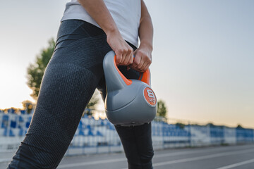Midsection of unknown caucasian woman female athlete holding kettlebell girya plastic weight while standing in stadium on track during training in summer evening copy space health and fitness concept